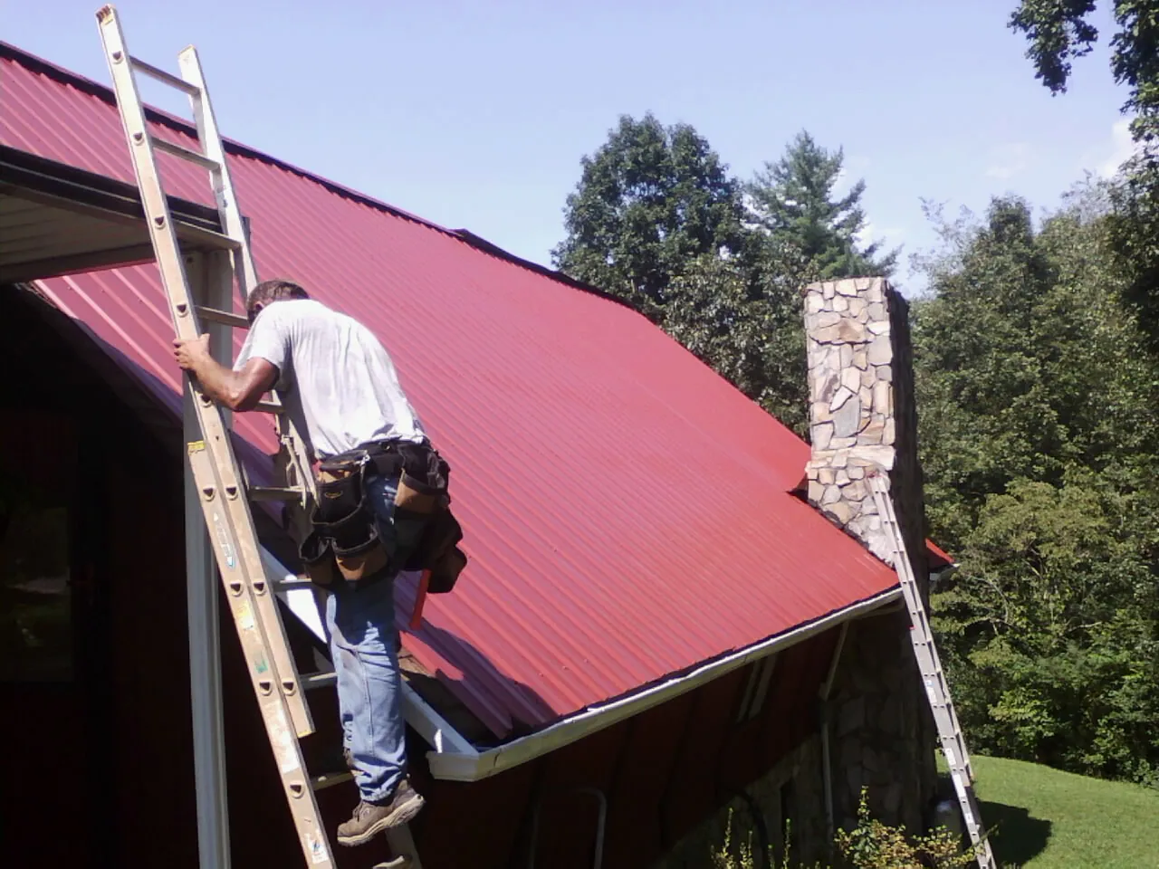 Roofer on a ladder beside a red metal roof and stone chimney — Soffit & Fascia Repair by Roofing Brothers in Anacortes